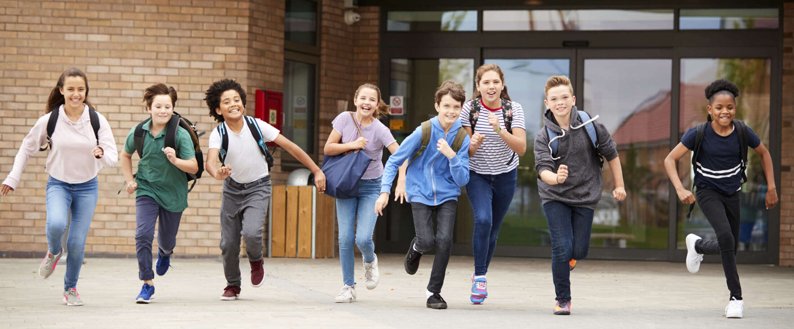 Group Of High School Students Running Out Of School Buildings Towards Camera At The End Of Class